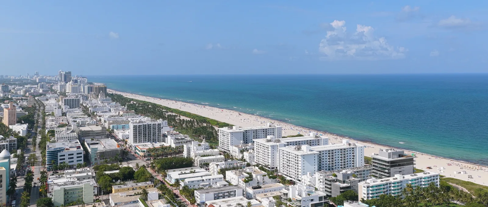 An aerial view along Miami Beach, white buildings and palms on the left and the sand and turquoise Atlantic running into the distance.