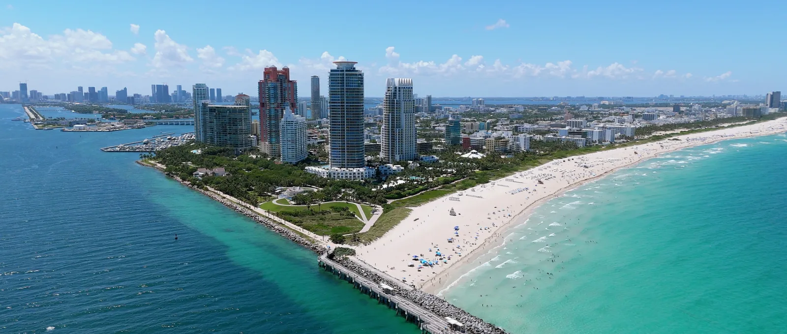 An aerial view of South Pointe at the southern tip of Miami Beach, towers behind a park, a fishing pier and rock jetty running into the water beside a white sand beach.